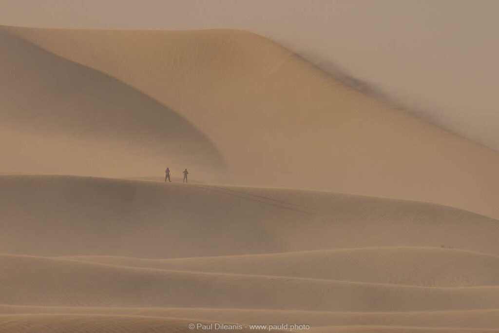 Lone figures brave a sandstorm in Death Valley dunes.
